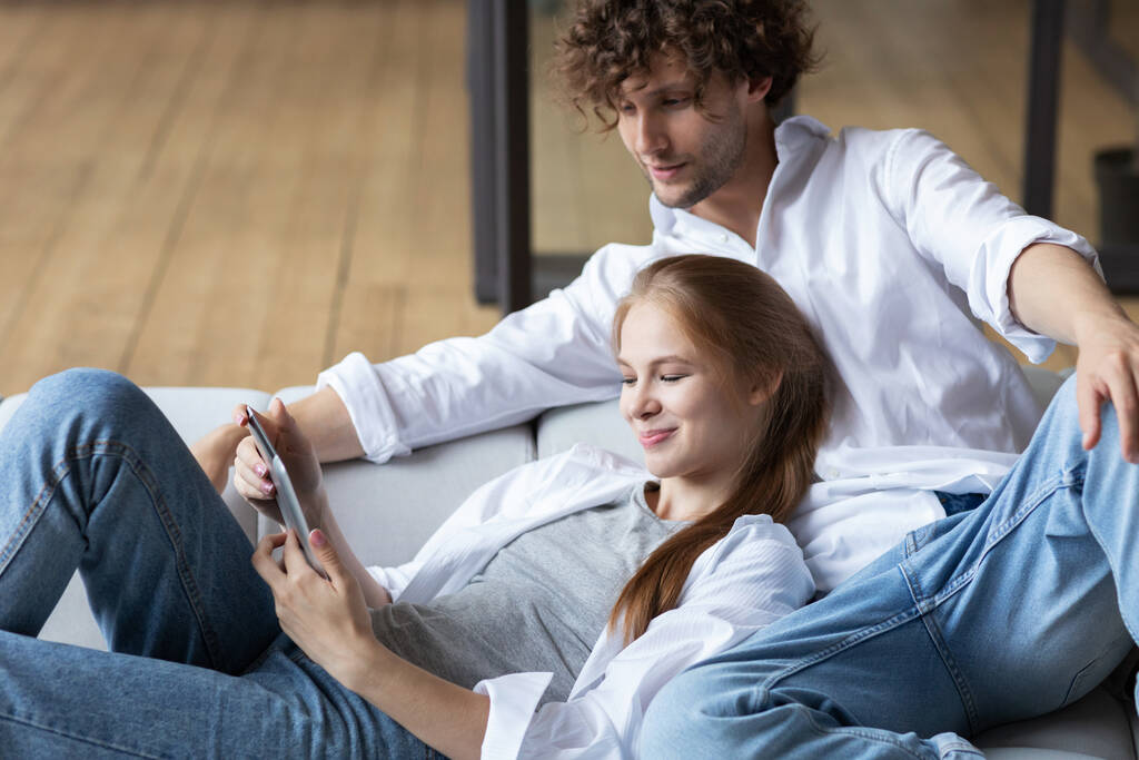 Szczęśliwa para korzystająca z tabletu cyfrowego podczas siedzenia na kanapie. Zakupy online. - Zdjęcie, obraz