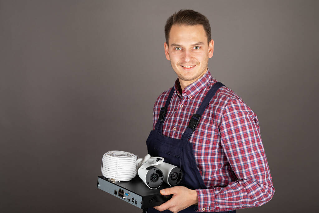 Ritratto di giovane allegro elettricista che tiene il kit della telecamera di sorveglianza davanti allo sfondo grigio - Foto, immagini