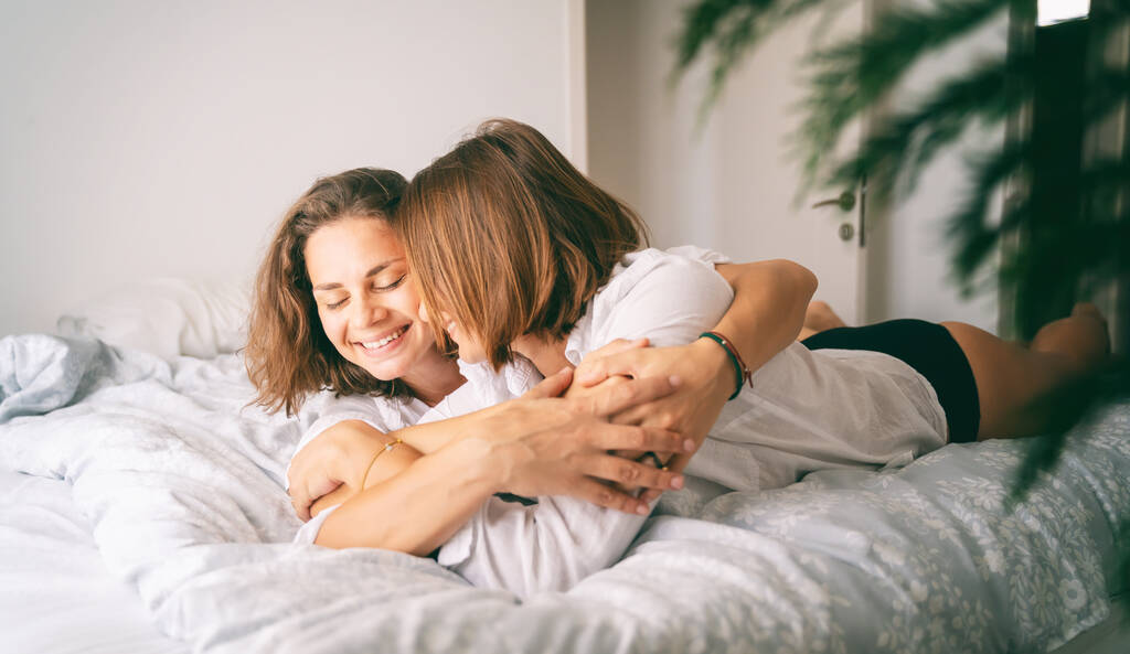 Young beautiful cheerful lesbian couple in love hugging while lying on bed at home - Photo, Image