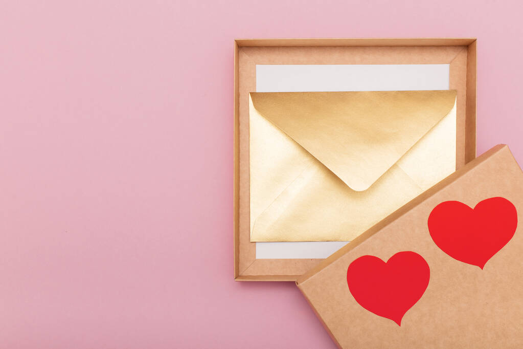 золотистий кольоровий паперовий конверт з червоним серцем на подарунковій коробці ізольований на рожевому фоні
 - Фото, зображення