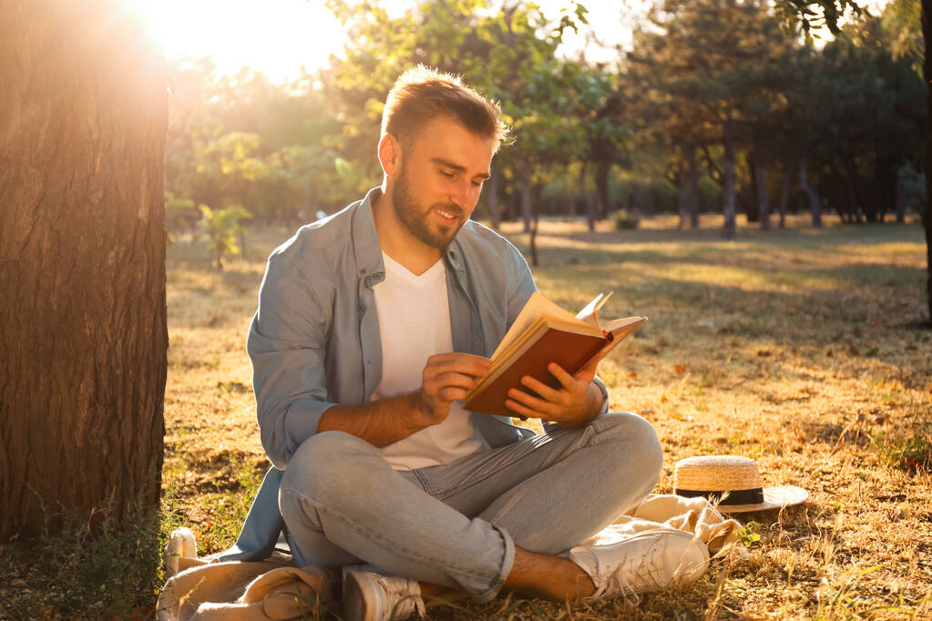 Молодой человек читает книгу на зеленой траве возле дерева в парке - Фото, изображение