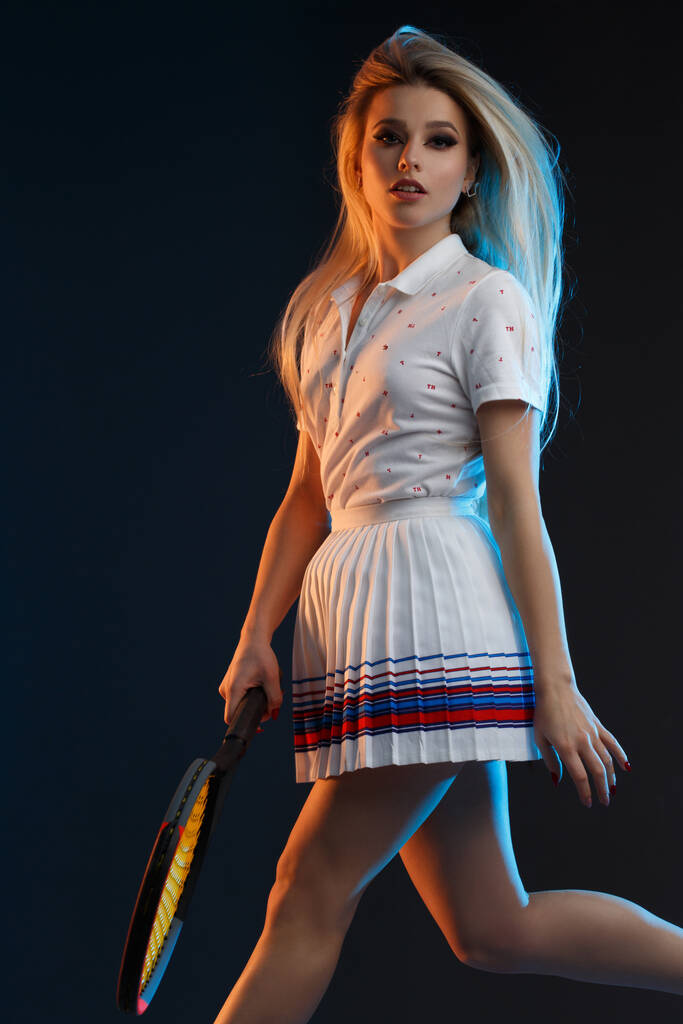 Bella atleta ragazza bionda con i capelli lunghi gioca a tennis su uno sfondo scuro con retroilluminazione gialla e blu. - Foto, immagini