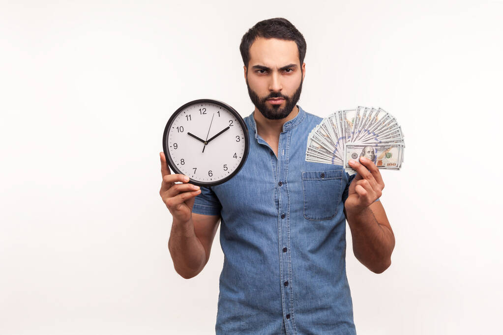 Assertive bossy bearded man holding big clock and dollars banknotes, looking at camera with aggressive expression, frustrated about hourly pay, debts. Indoor studio shot isolated on white background - Photo, Image