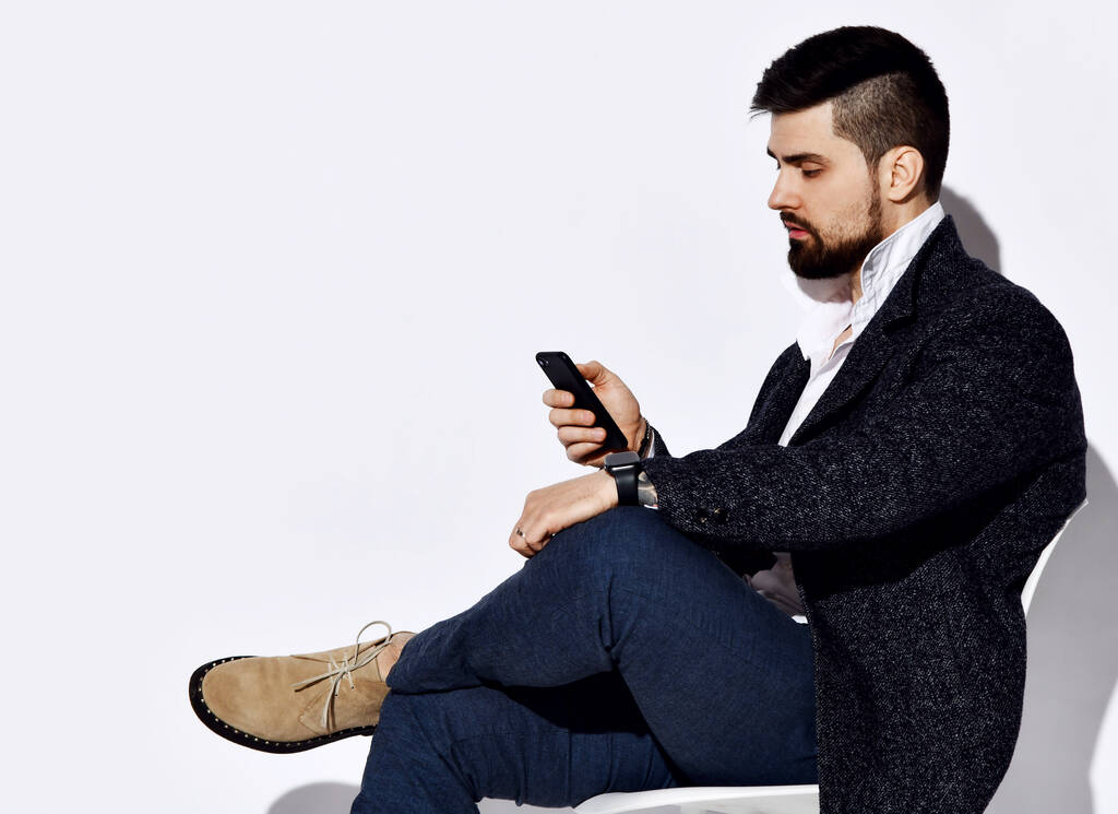  Brutal hombre barbudo en jeans y elegante chaqueta de tweed casual está utilizando su teléfono inteligente de mensajería noticias de lectura. Vista lateral - Foto, Imagen