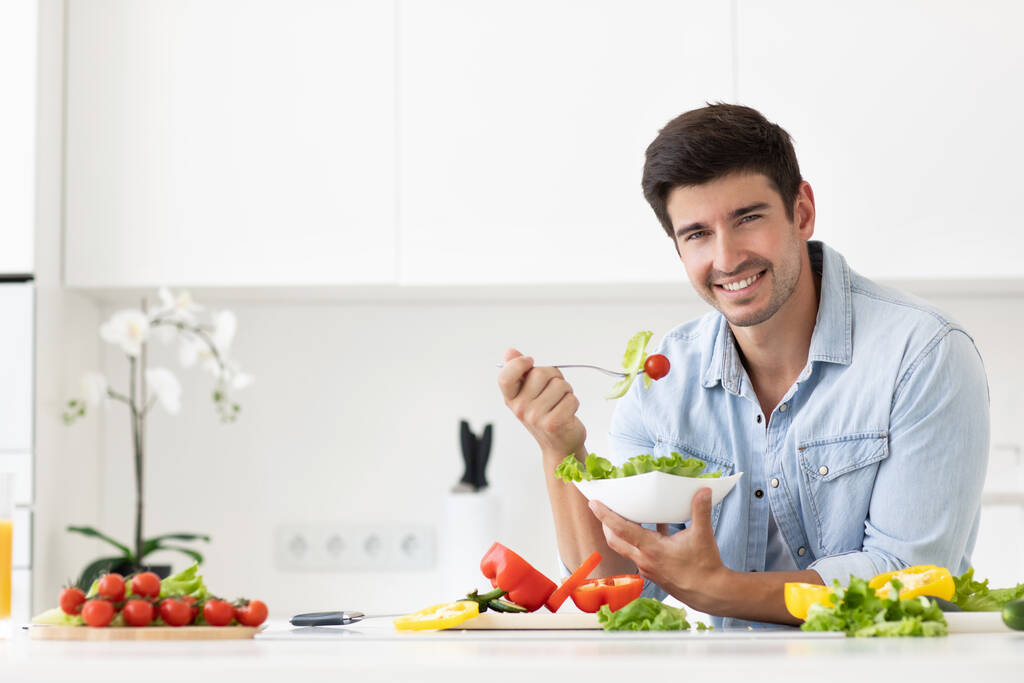 Komea mies syö salaattia tuoreiden vihannesten kanssa keittiössä. Terveellinen ja vegaani ruoka käsite. - Valokuva, kuva