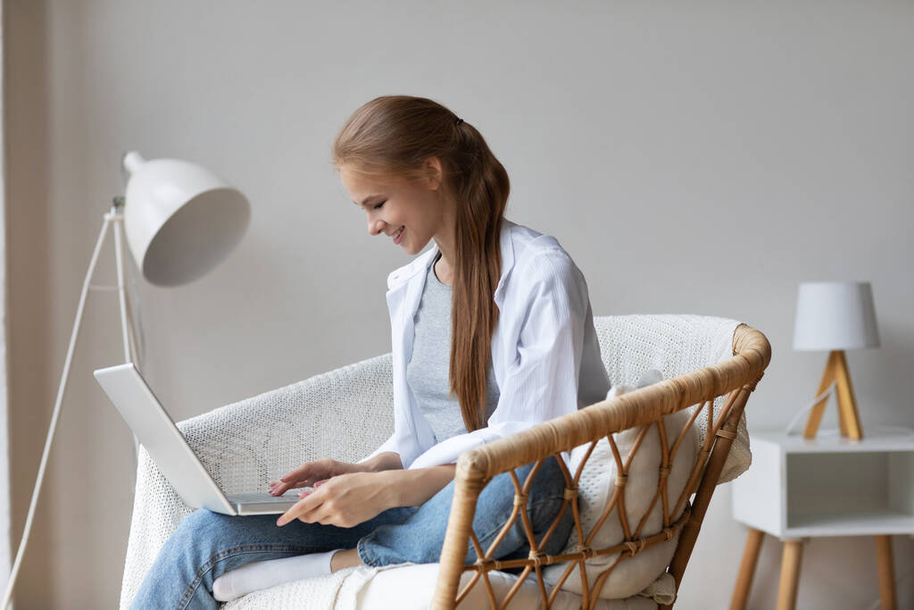 Keyifli, rahat bir kadın evdeki bir sandalyede otururken dizüstü bilgisayar kullanıyor. Uzaktan iletişim. - Fotoğraf, Görsel