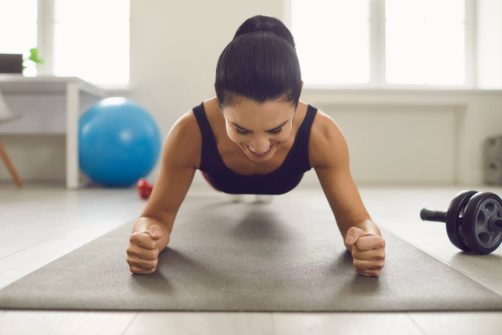 Сосредоточенная улыбающаяся женщина держит доску и делает упражнения на мышцах живота в ярком спортзале. - Фото, изображение