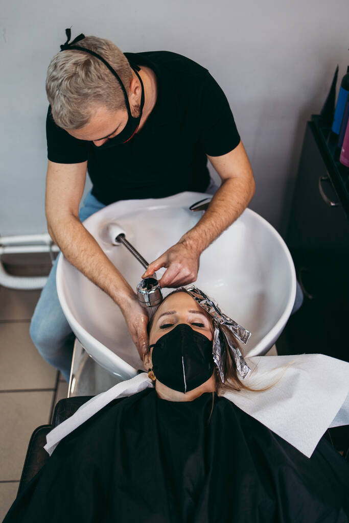 Cabeleireiro lavar o cabelo de uma bela jovem mulher adulta no salão de cabeleireiro. Ela está usando máscara protetora como proteção contra pandemia de vírus. - Foto, Imagem