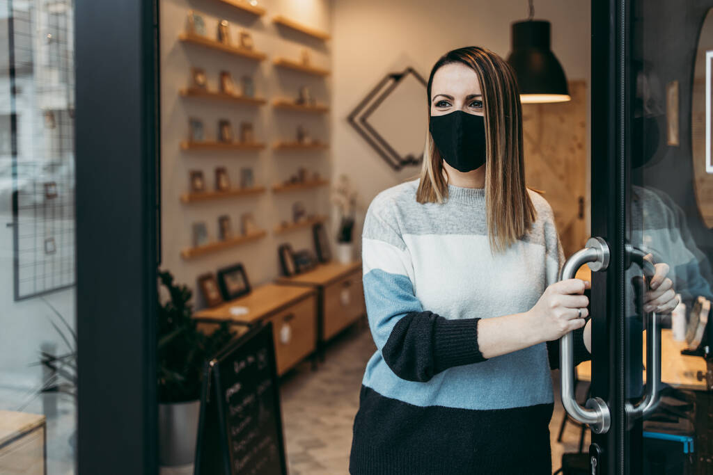 Fiatal és vonzó női művész arcvédő maszkkal áll a nyitott műhely ajtaján és néz ki. Magabiztos és komoly. Covid-19 nyitott üzleti koncepció. - Fotó, kép
