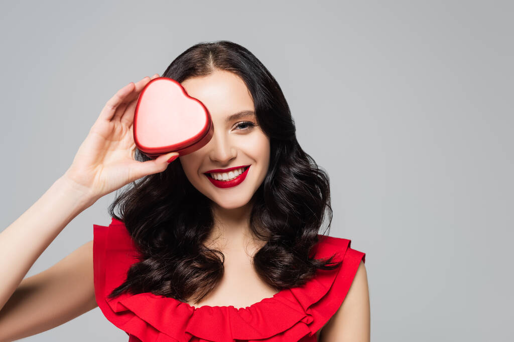 mujer alegre con labios rojos que cubre el ojo mientras sostiene la caja de regalo en forma de corazón aislada en gris - Foto, imagen