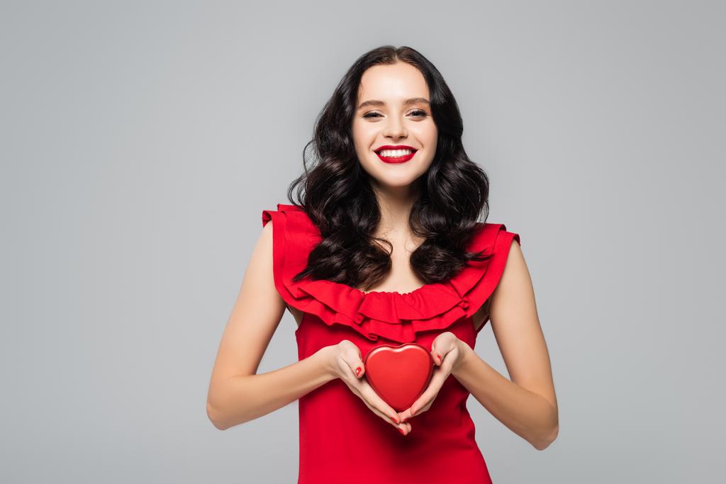 glimlachende vrouw met rode lippen houden hartvormige geschenkdoos geïsoleerd op grijs - Foto, afbeelding