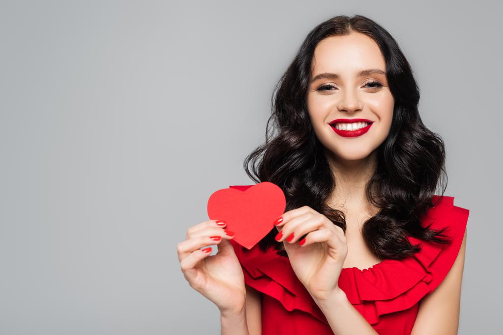 Ευτυχισμένη νεαρή γυναίκα που κρατά κόκκινη χάρτινη καρδιά απομονωμένη στο γκρι - Φωτογραφία, εικόνα