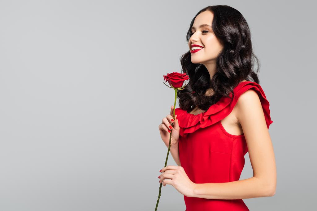 χαμογελαστή νεαρή γυναίκα που κρατά το κόκκινο τριαντάφυλλο απομονωμένο στο γκρι  - Φωτογραφία, εικόνα