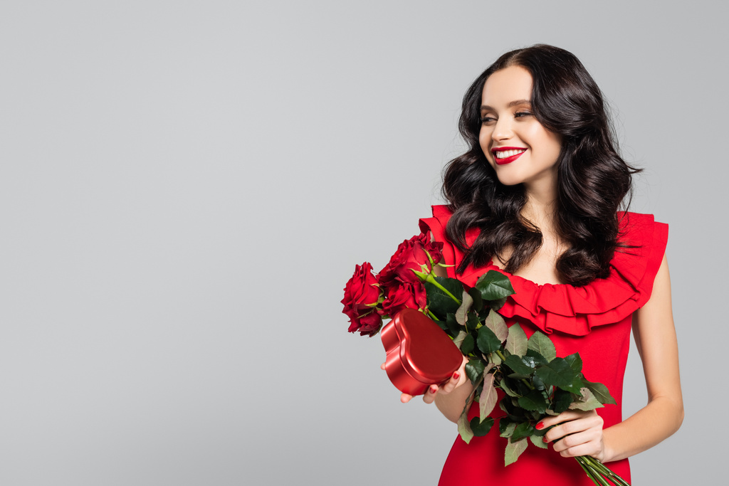 happy young woman holding bouquet of red roses and heart-shaped box isolated on grey  - Photo, Image
