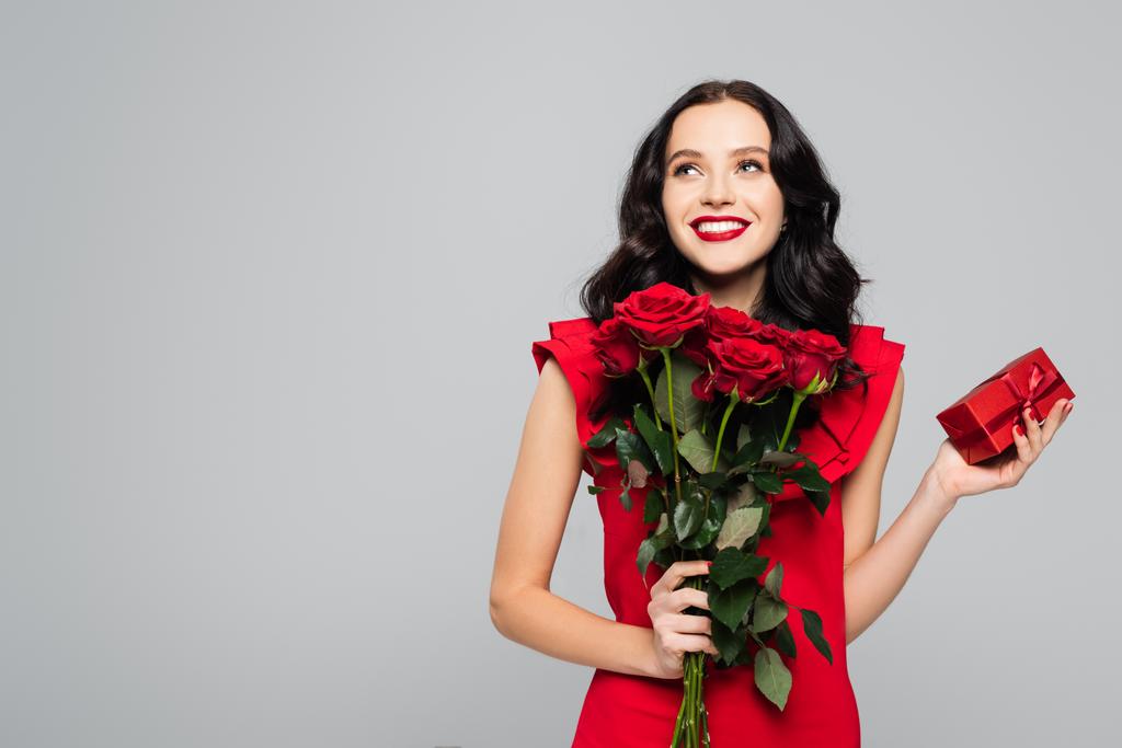 ευχαριστημένη γυναίκα κρατώντας τριαντάφυλλα και τυλιγμένο κουτί απομονώνονται σε γκρι  - Φωτογραφία, εικόνα