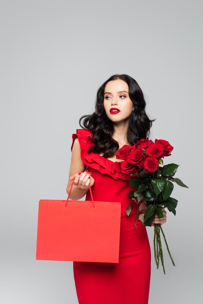 ショッピングバッグとバラを持つブルネットの女性は灰色で隔離された - 写真・画像