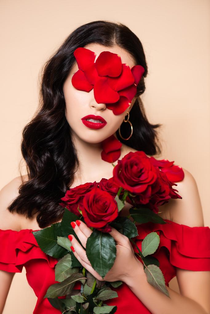 mujer joven con pétalos en la cara sosteniendo rosas rojas aisladas en rosa - Foto, Imagen