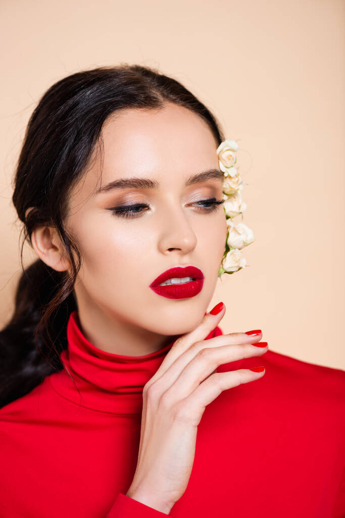 femme sensuelle avec des lèvres rouges et des fleurs blanches sur le visage regardant loin isolé sur rose  - Photo, image