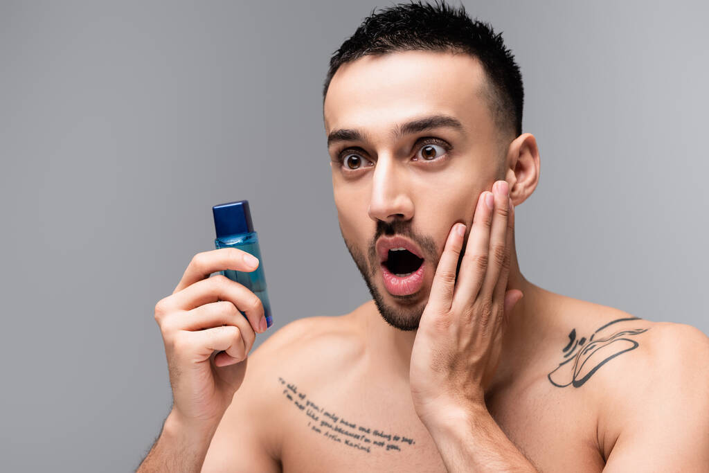 geschokt latino man aanraken gezicht terwijl het houden van fles aftershave lotion geïsoleerd op grijs - Foto, afbeelding