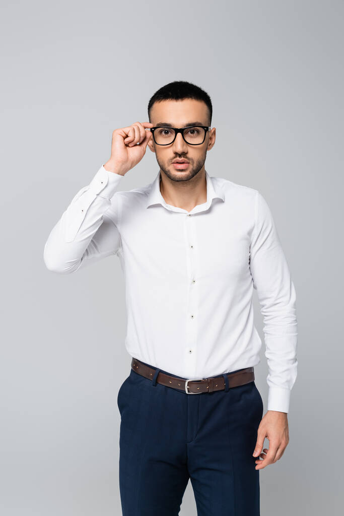 brunette hispanic businessman adjusting eyeglasses while looking at camera isolated on grey - Photo, Image