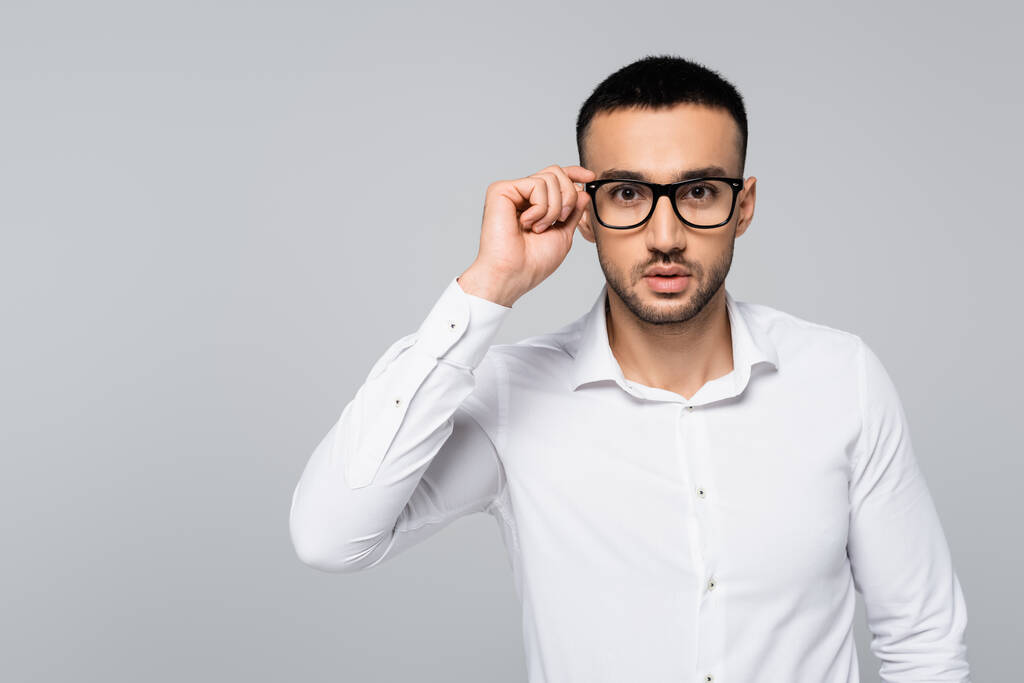 νεαρός ισπανόφωνος διευθυντής σε λευκό πουκάμισο προσαρμογή γυαλιά ενώ κοιτάζοντας κάμερα που απομονώνονται σε γκρι - Φωτογραφία, εικόνα
