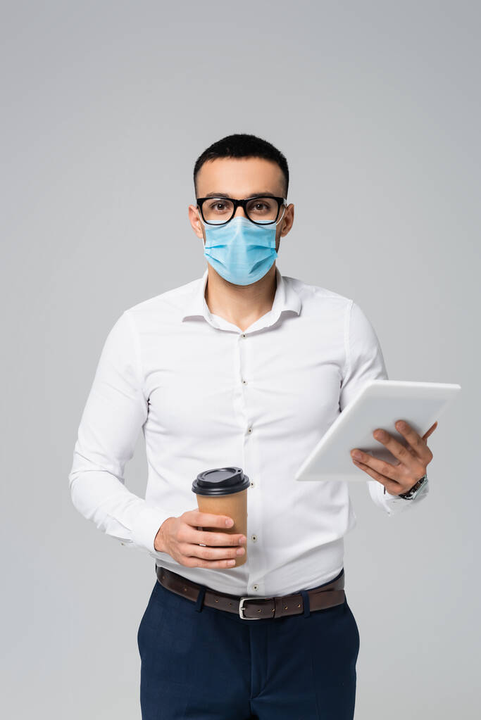 young hispanic businessman in medical mask holding digital tablet and coffee to go isolated on grey - Photo, Image
