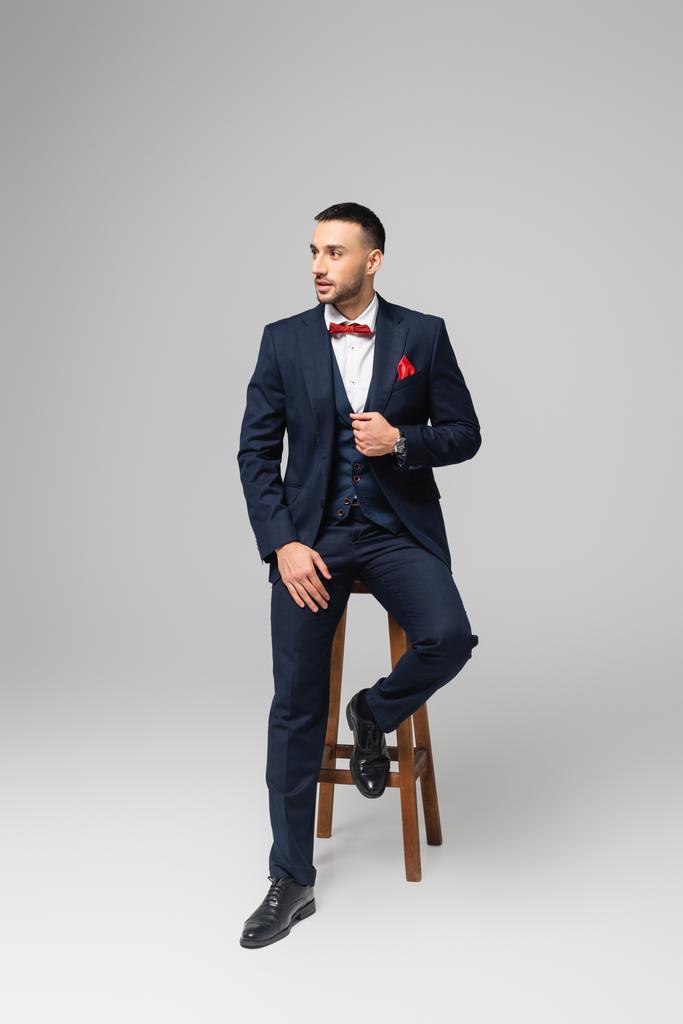 plný pohled na mladého hispánského muže v elegantním obleku pózuje na vysoké stoličce na šedé - Fotografie, Obrázek