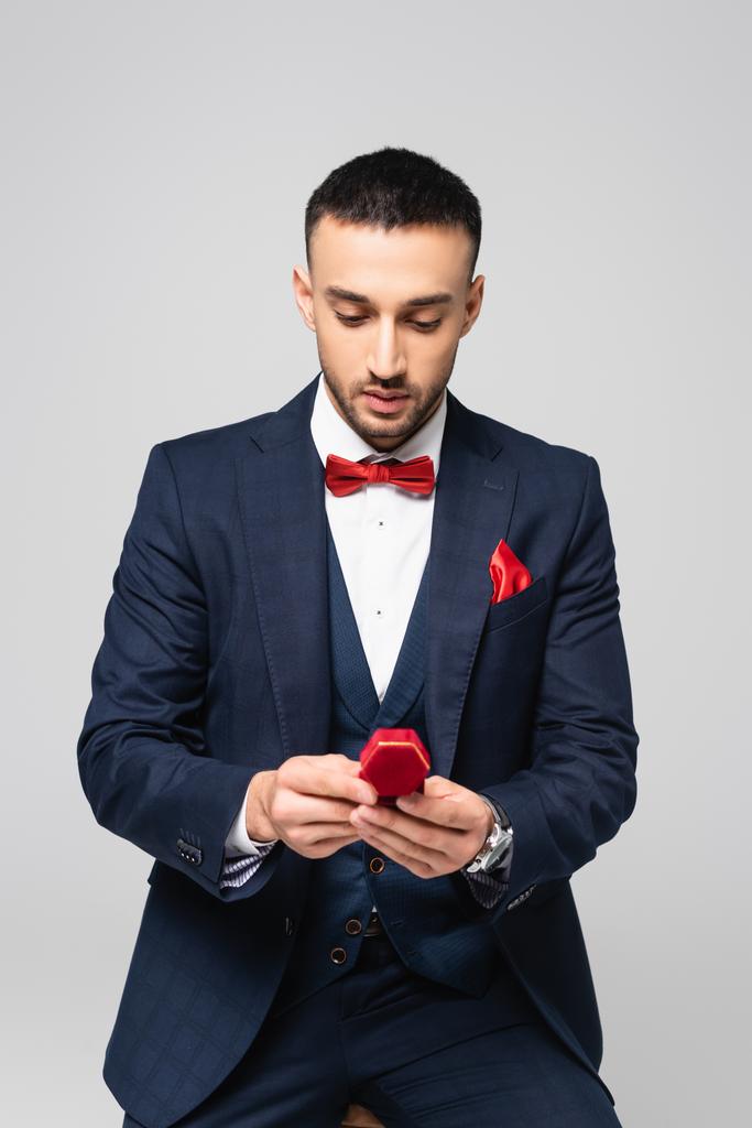 κομψός ισπανόφωνος άντρας με μπλε κοστούμι κοιτάζει την κόκκινη κοσμηματοθήκη που είναι απομονωμένη στο γκρι - Φωτογραφία, εικόνα