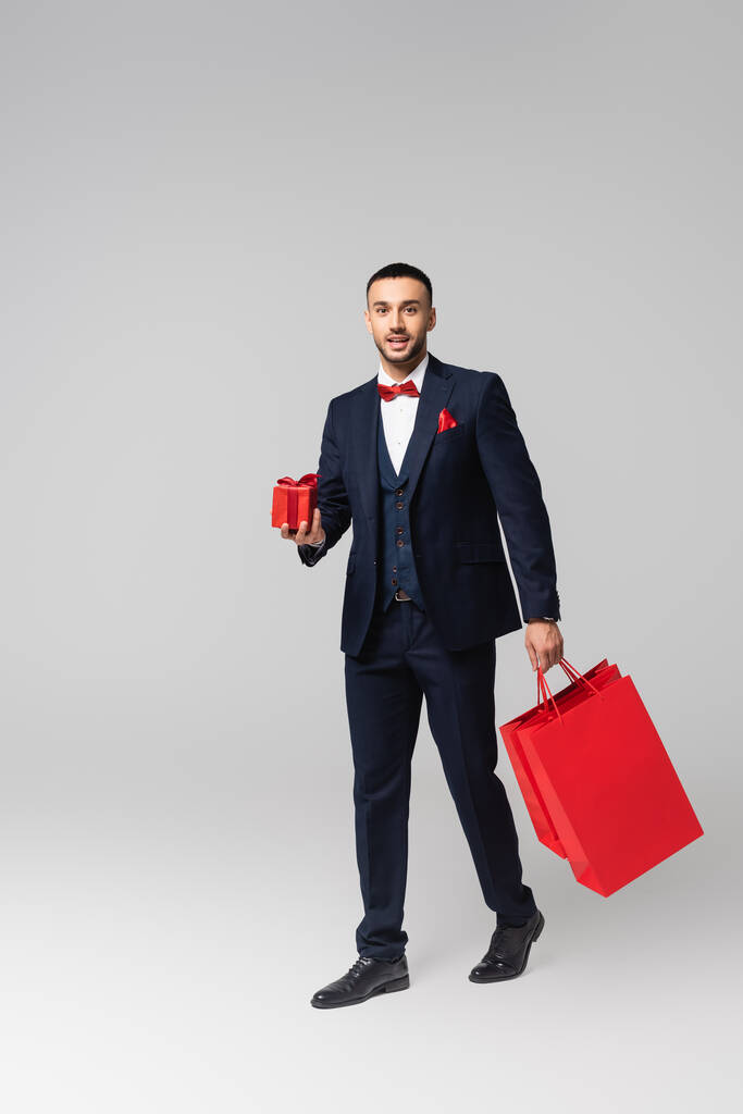молодой элегантный латиноамериканец ходит с красной подарочной коробкой и сумками для покупок на сером - Фото, изображение
