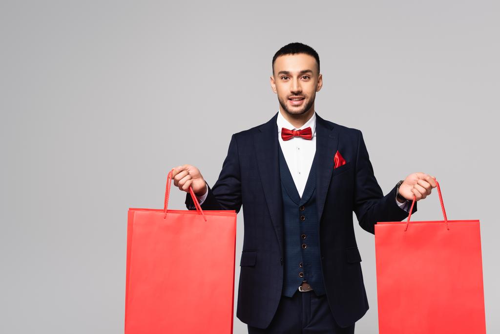 happy hispanic man in elegant suit holding red shopping bags isolated on grey - Photo, Image