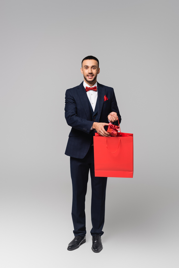täyspitkä näkymä onnellinen latino mies tyylikäs puku laittaa läsnä ostoskassi harmaa - Valokuva, kuva