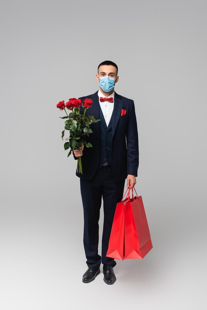 täyspitkä näkymä tyylikäs latino mies lääketieteellinen naamio punaisia ruusuja ja ostoskassit harmaa - Valokuva, kuva