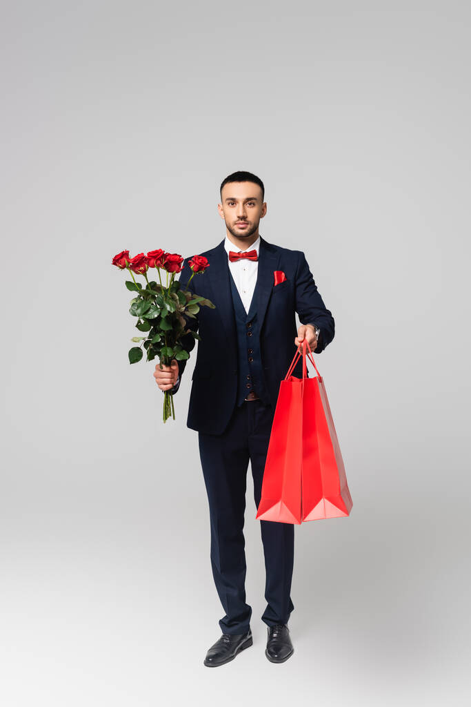 vista completa del elegante hombre hispano sosteniendo bolsas de compras rojas y rosas rojas en gris - Foto, imagen