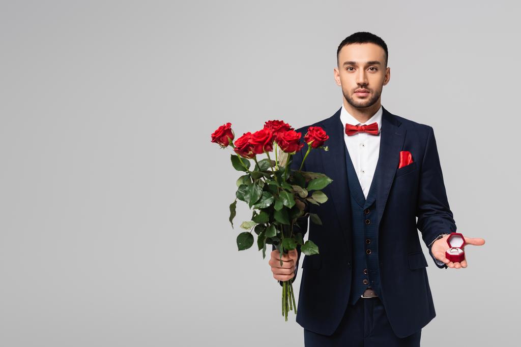elegantti latino mies sininen puku tilalla punaisia ruusuja ja koruja laatikko eristetty harmaa - Valokuva, kuva