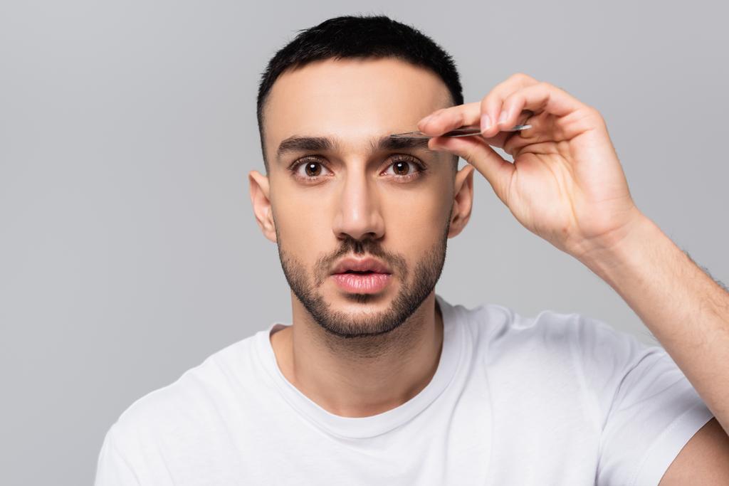 brunette hispanique homme regardant caméra tout en épilant sourcils isolés sur gris - Photo, image