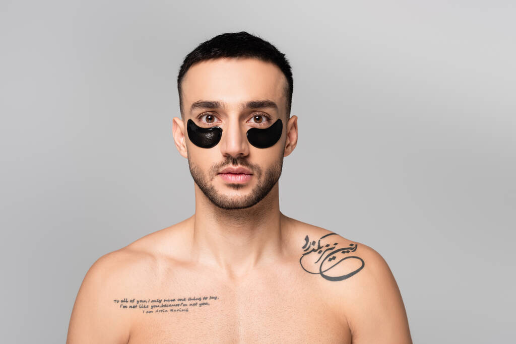 без рубашки, татуированный латиноамериканец с повязками на глазу, смотрящий на камеру, изолированную на сером - Фото, изображение