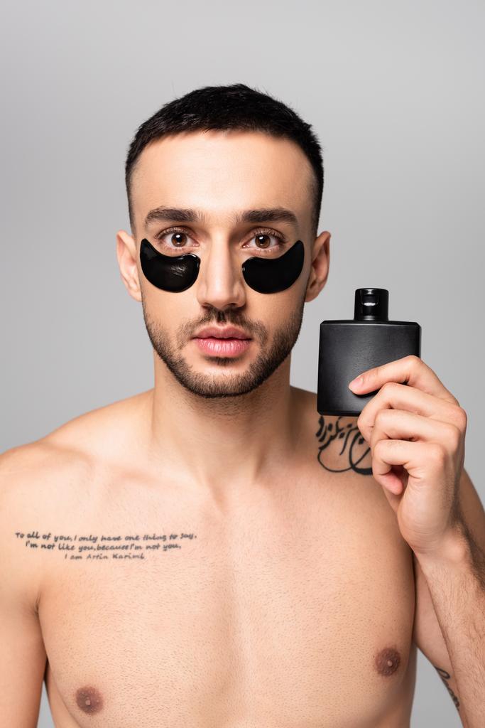 Üzerinde göz bandı olan tişörtsüz İspanyol adam gri renkte bir şişe parfüm suyu tutuyor. - Fotoğraf, Görsel