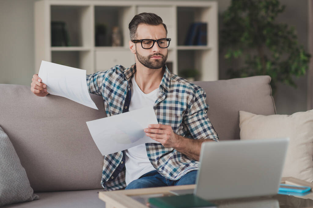 Zdjęcie młody przystojny mężczyzna skoncentrowane poważne analizować papier dokument wygląd laptop siedzieć kanapa domu odległość praca - Zdjęcie, obraz