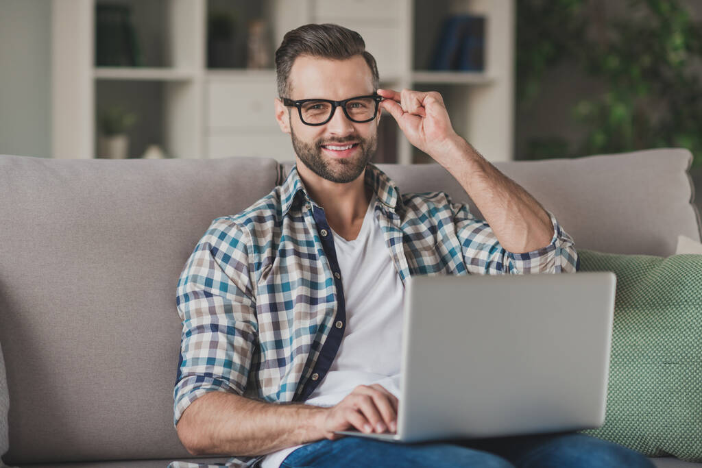 Zdjęcie młodego przystojnego biznesmena szczęśliwy pozytywny uśmiech siedzieć sofa dom odległość praca przeglądać laptop ręka dotyk okulary - Zdjęcie, obraz