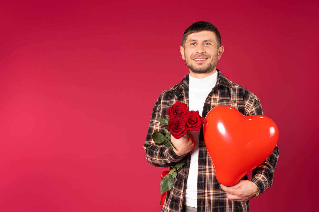 Człowiek z bukietem czerwonych róż i czerwonym balonem w kształcie serca na czerwonym tle z przestrzenią boczną. Cupidon w Walentynki - Zdjęcie, obraz