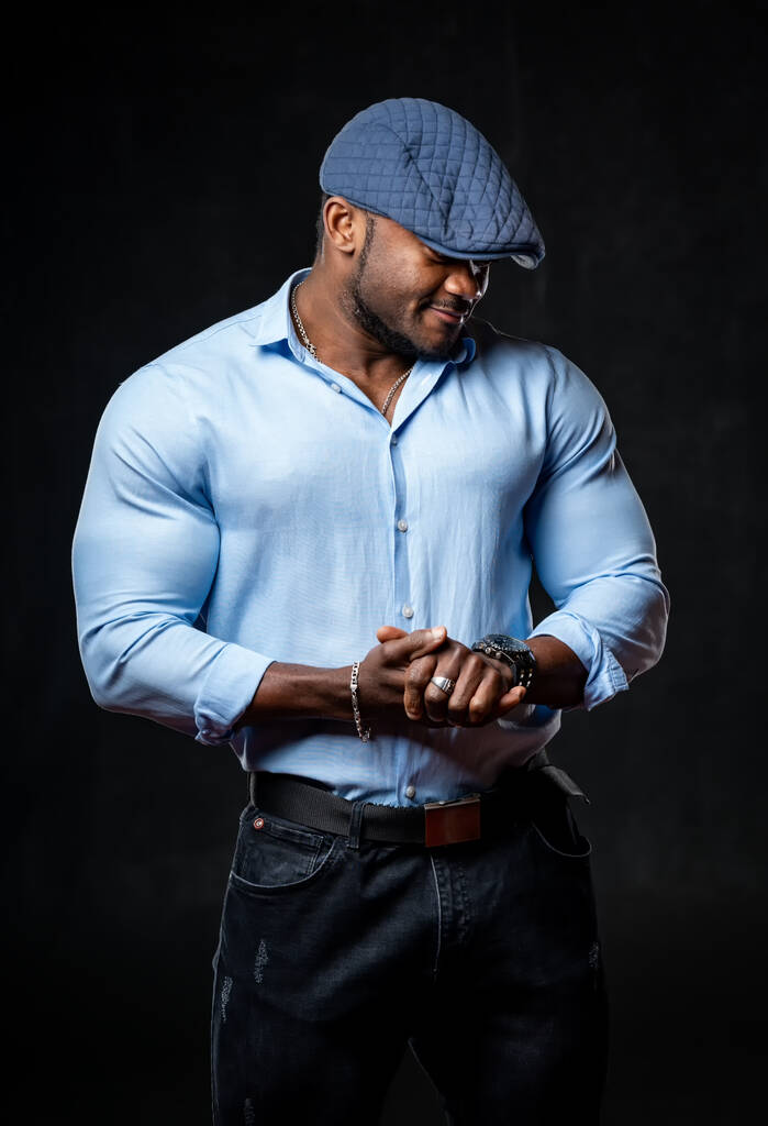 Afroamerikaner mit blauer Mütze, die das halbe Gesicht bedeckt, posiert in Jeans und blauem Hemd vor der Kamera. Schwarzer Hintergrund. - Foto, Bild