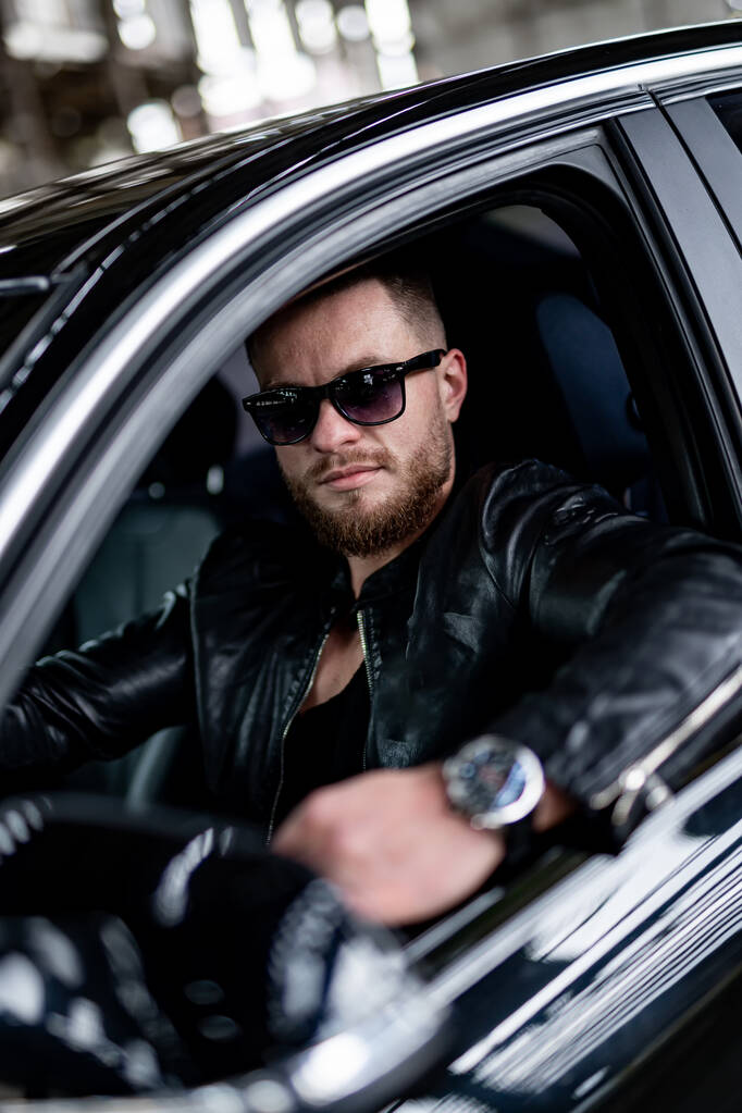 Brutaler Mann in Lederjacke sitzt im Luxusauto. Teure Uhren auf der Hand eines gutaussehenden Mannes. - Foto, Bild