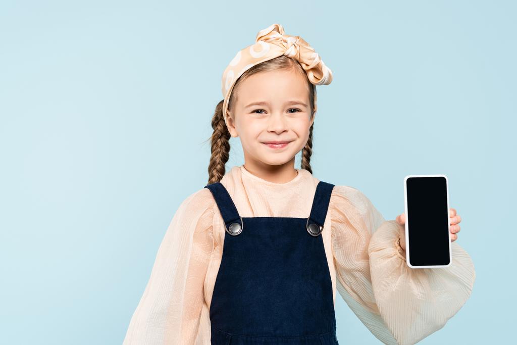 niño feliz con coletas sosteniendo teléfono inteligente con pantalla en blanco aislado en azul - Foto, Imagen