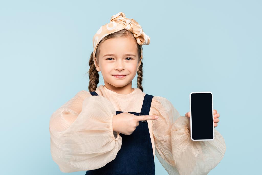 niña con coletas apuntando con el dedo al teléfono inteligente con pantalla en blanco aislado en azul - Foto, imagen