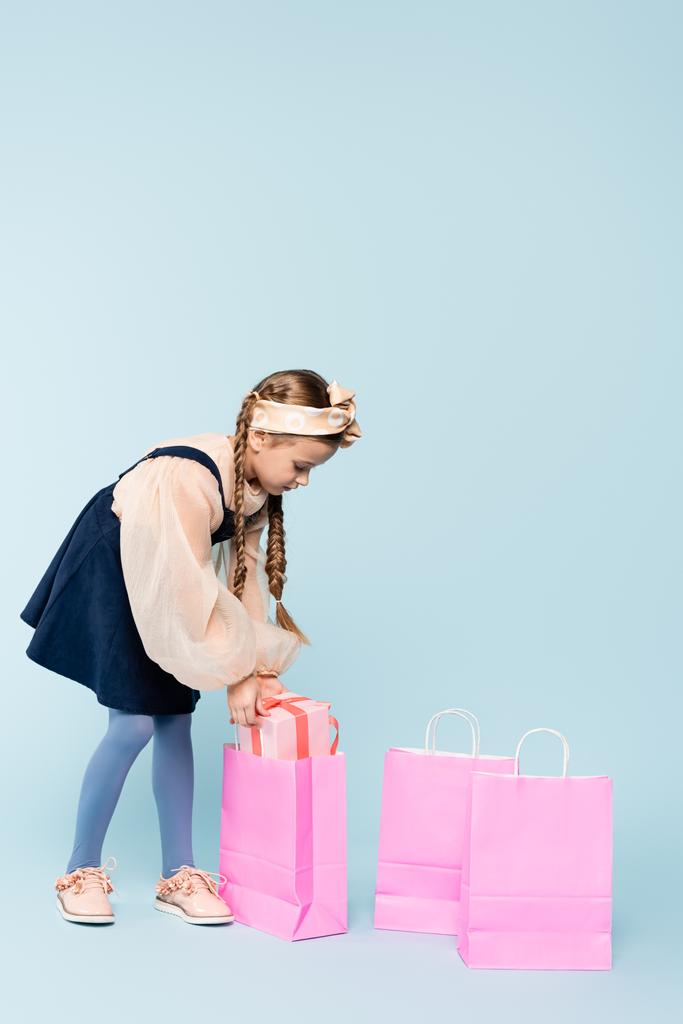 full length of little girl in dress putting present in shopping bag on blue - Photo, Image