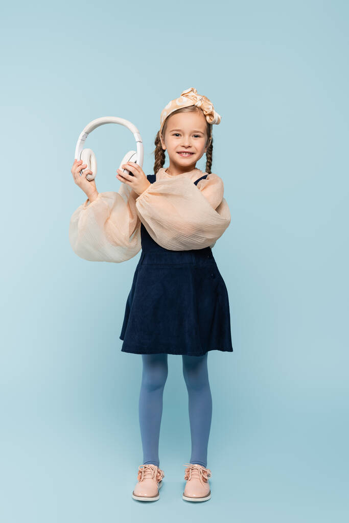повна довжина веселого дитини в пов'язці з бантом, що тримає бездротові навушники на синьому
  - Фото, зображення