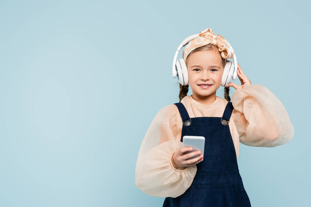 niño alegre en diadema con arco ajustando auriculares inalámbricos mientras mantiene el teléfono inteligente aislado en azul  - Foto, imagen