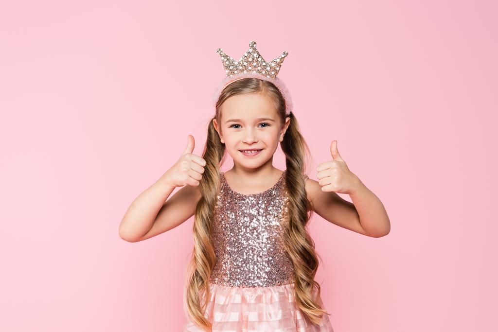alegre niña en vestido y corona mostrando los pulgares hacia arriba aislado en rosa  - Foto, Imagen