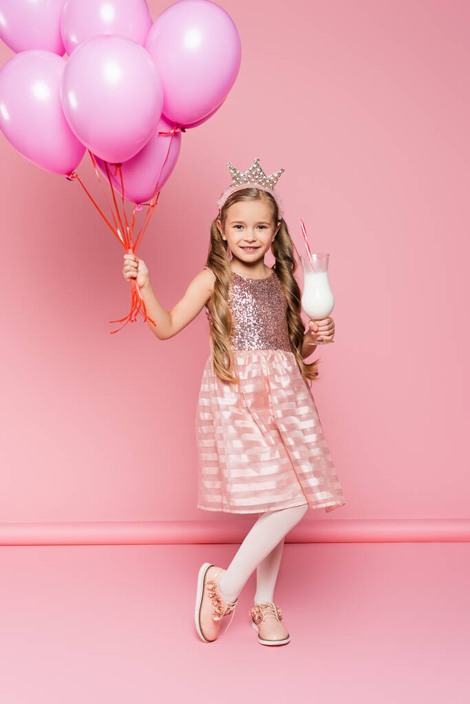 πλήρες μήκος του χαρούμενου μικρού κοριτσιού σε φόρεμα και στέμμα κρατώντας γυαλί με milkshake και μπαλόνια σε ροζ  - Φωτογραφία, εικόνα