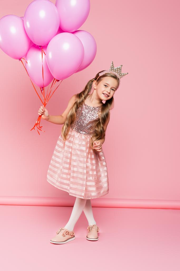 full length of cheerful little girl in dress and crown holding balloons on pink  - Photo, Image